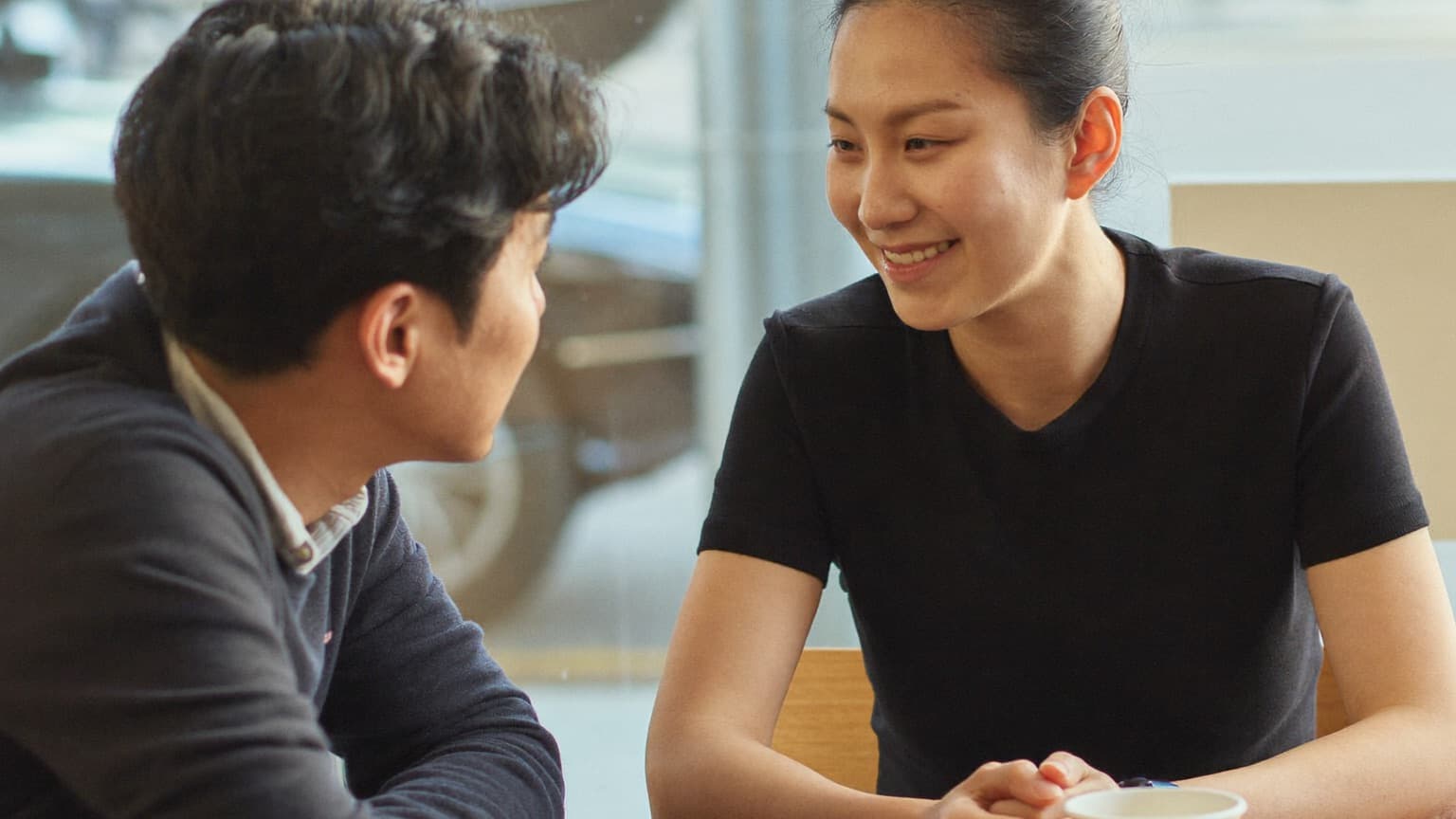 Two people in their late twenties sitting across a small table in a bright cafe, one leaning forward mid-question, the other listening and smiling.